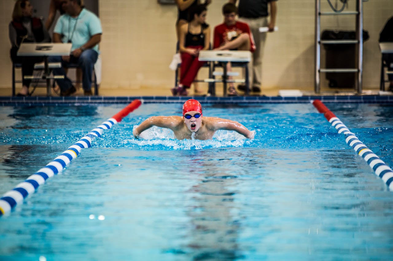 A young swimmer in a red cap competes in a butterfly stroke during an indoor pool race.