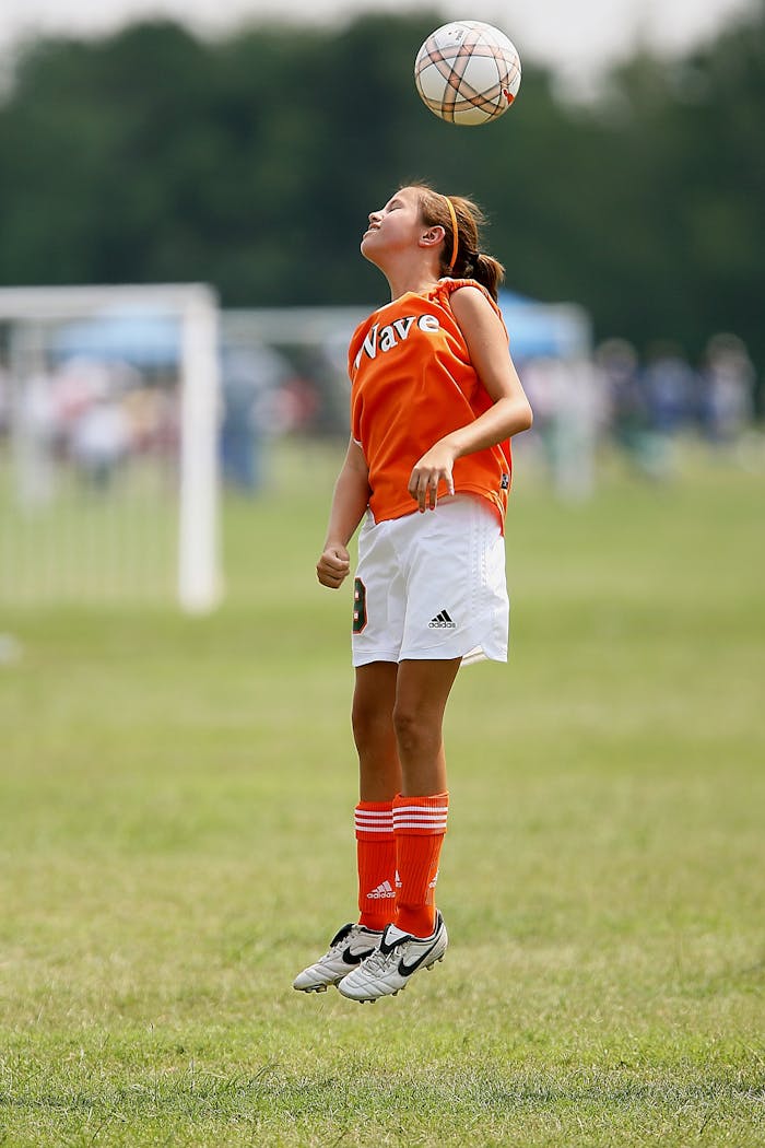 A young girl jumps to head a soccer ball during a game on a grassy field.